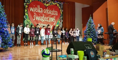 Na zdjęciu wolontariusze Wielkiej Orkiestry Świątecznej Pomocy na scenie sali widowiskowej w Dusznikach na tle logo Orkiestry.