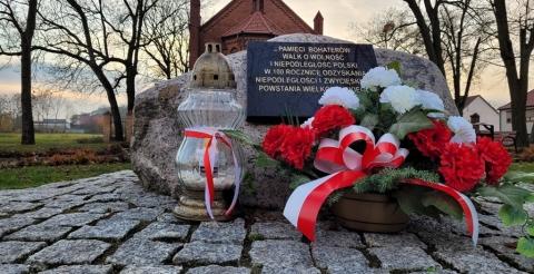 Tablica pamiątkowa ku pamięci Powstańców Wielkopolskich udekorowana kwiatami i zniczem.