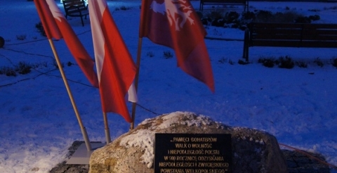Uroczystość 103. rocznicy wybuchu Powstania Wielkopolskiego pod tablicą pamiątkową na skwerze przy ul. Powstańców Wielkopolskich. Na zdjęciu tablica pamiątkowa z wiązankami kwiatów, w tle dwie flagi powstańcze i flaga narodowa oraz budynek kościoła oświetlony biało-czerwoną iluminacją.