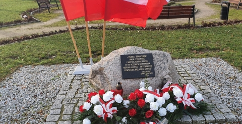 Na zdjęciu pamiątkowa tablica udekorowana złożonymi wiązankami. 