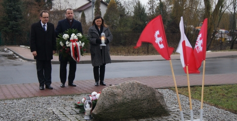 Na zdjęciu (od lewej) Wójt Gminy Duszniki Roman Boguś, Wojewoda Wielkopolski Michał Zieliński i Starosta Szamotulski Beata Hanyżak stoją przed tablicą upamiętniającą Powstańców Wielkopolskich. Wojewoda trzyma biało-czerwony wieniec kwiatów, Starosta trzyma zapalony znicz. Po prawej stronie zdjęcia, na pierwszym planie, powiewają flagi: biało-czerwona i dwie powstańcze. 