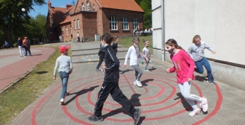 Na zdjęciu uczniowie biegają po liniach narysowanych na chodniku przed szkołą.