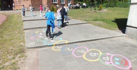 Na zdjęciu uczniowie chodzą po okręgach narysowanych na chodniku przed szkołą.