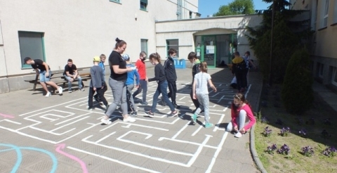 Na zdjęciu uczniowie z nauczycielem chodzą po liniach narysowanych na chodniku przed szkołą.