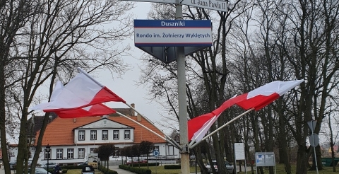 Na zdjęciu udekorowany flagami państwowymi słup znajdujący się na środku głównego ronda w Dusznikach z oznaczeniem nazwy Rondo im. Żołnierzy Wyklętych