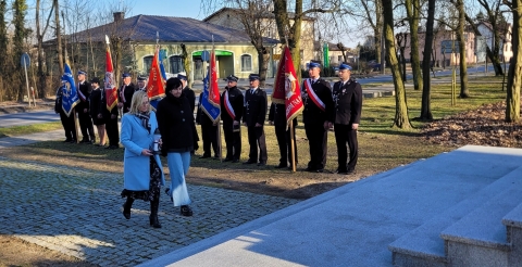 Delegacja ze zniczem zmierza w kierunku pomnika. W tle poczty sztandarowe Ochotniczych Straży Pożarnych ze sztandarami.