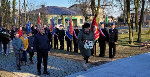 Delegacja z wiązanką biało-czerwonych kwiatów zmierza w kierunku pomnika. W tle poczty sztandarowe Ochotniczych Straży Pożarnych ze sztandarami.