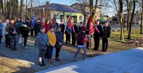 Delegacja dzieci szkolnych z wiązanką biało-czerwonych kwiatów zmierza w kierunku pomnika. W tle poczty sztandarowe Ochotniczych Straży Pożarnych ze sztandarami.
