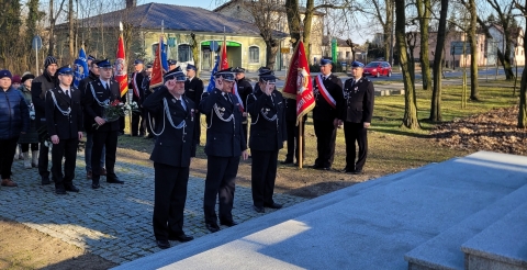 Trzyosobowa delegacja Zarządu Gminnego Ochotniczych Straży Pożarnych salutuje w kierunku pomnika. W tle poczty sztandarowe Ochotniczych Straży Pożarnych ze sztandarami.