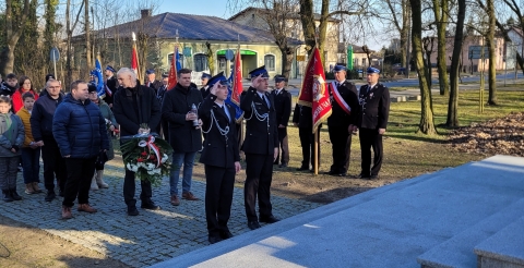Dwuosobowa delegacja Ochotniczych Straży Pożarnych salutuje w kierunku pomnika. W tle poczty sztandarowe Ochotniczych Straży Pożarnych ze sztandarami.