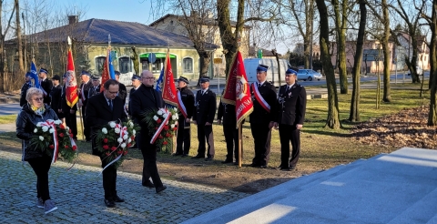 Delegacja samorządu gminy z wiązankami biało-czerwonych kwiatów zmierza w kierunku pomnika. W tle poczty sztandarowe Ochotniczych Straży Pożarnych ze sztandarami.