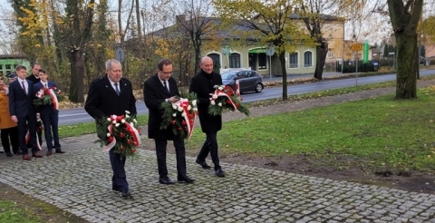 Wójt z radnym i sołtysem z wiązankami kwiatów zmierzają w kierunku pomnika. W tle pozostałe delegacje.