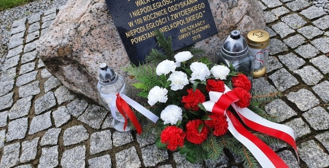 Na zdjęciu tablica przy skwerze przy ulicy Powstańców Wielkopolskich w Dusznikach przyozdobiona wiązanką biało-czerwonych kwiatów i zniczami.